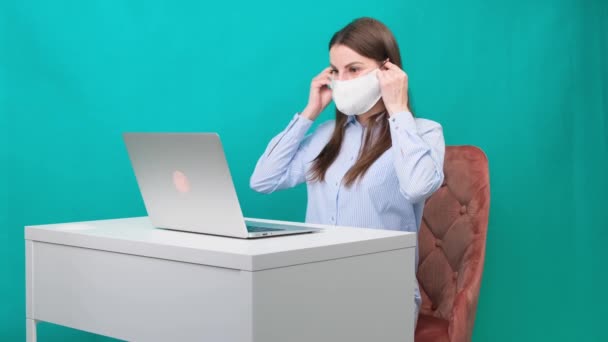 Vrouw zet een beschermend masker op tijdens het werken op een laptop op de werkplek of thuis tijdens een pandemie. Het begrip werk tijdens quarantaine en zelfisolatie. — Stockvideo