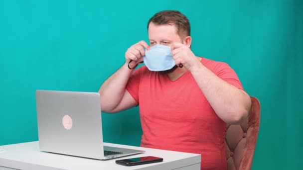 Een man met een baard in een roze T-shirt zet een beschermend medisch masker op voor het thuis werken op een laptop op een groene achtergrond. Quarantaineconcept — Stockvideo