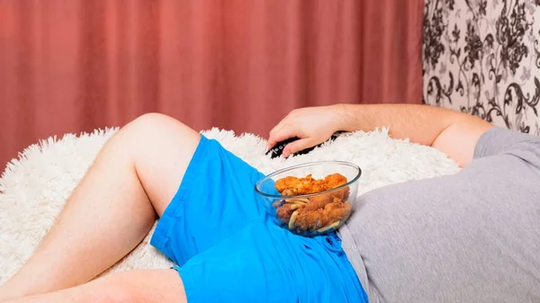 Dikke man met een grote buik ligt op de bank met fast food en verveelt zich voor de tv met de afstandsbediening in zijn hand, close-up. — Stockfoto