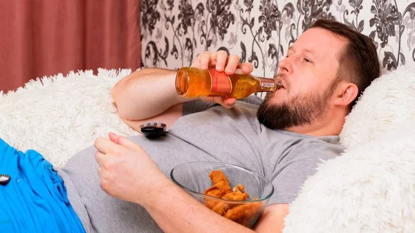 Barbudo gordo con un gran vientre se encuentra en el sofá con comida rápida, bebe cerveza y echa de menos el televisor con el mando a distancia en la mano, primer plano . — Foto de Stock