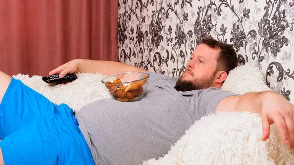 Жирний чоловік з великим животом лежить на дивані з фаст-фудом і нудно перед телевізором з пультом в руці, крупним планом . — стокове фото