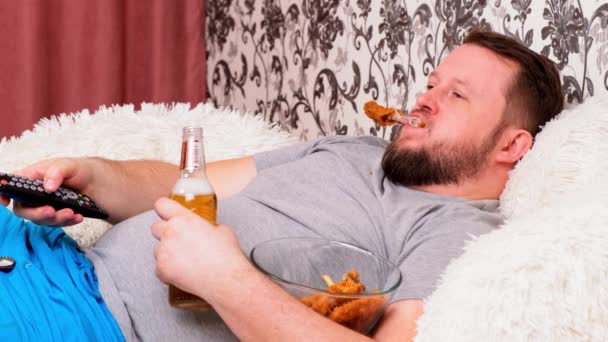 Uomo grasso barbuto con una grande pancia si trova sul divano con fast food, beve birra e manca la TV con il telecomando in mano, primo piano . — Video Stock