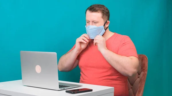 Ein bärtiger Mann im rosafarbenen T-Shirt setzt sich eine medizinische Schutzmaske auf, um zu Hause vor grünem Hintergrund am Laptop zu arbeiten. Quarantäne-Konzept — Stockfoto