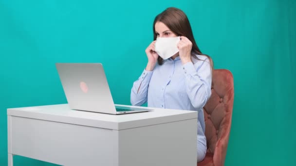 Vrouw zet een beschermend masker op tijdens het werken op een laptop op de werkplek of thuis tijdens een pandemie. Het begrip werk tijdens quarantaine en zelfisolatie. — Stockvideo