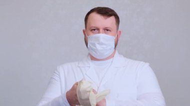 Koruyucu tıbbi maskeli beyaz elbiseli sakallı adam ellerine eldiven takıyor.