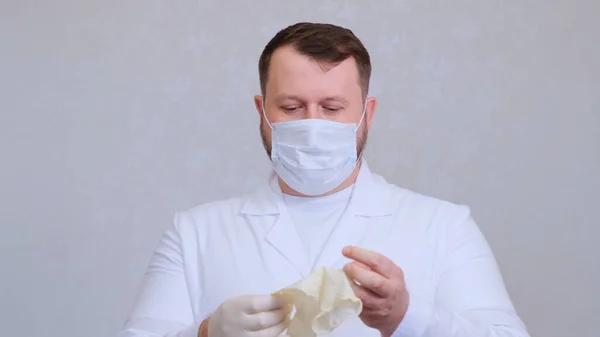 Homem barbudo em uma máscara médica protetora em roupas brancas coloca luvas em suas mãos — Fotografia de Stock