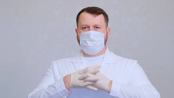 Homem barbudo em uma máscara médica protetora em roupas brancas coloca luvas em suas mãos — Fotografia de Stock