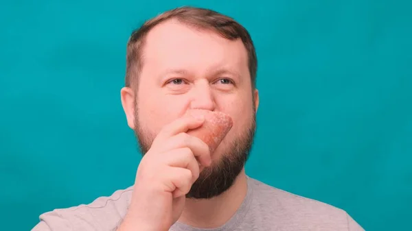 Bearded man bites eating a big salami sausage on a green background. Diet concept failed, willpower in nutrition, junk food — Stock Photo, Image