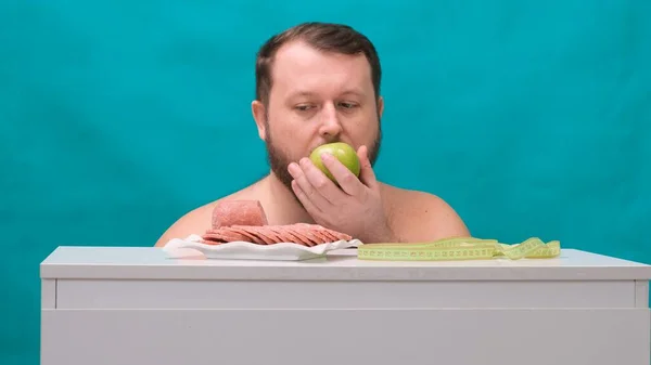 Homem gordo barbudo escolhe entre uma salsicha ou uma maçã verde em uma tela verde. Um homem triste morde uma maçã verde. Conceito Resistência à tentação, fast food, alimentação saudável, dieta, cuidados com o corpo . — Fotografia de Stock