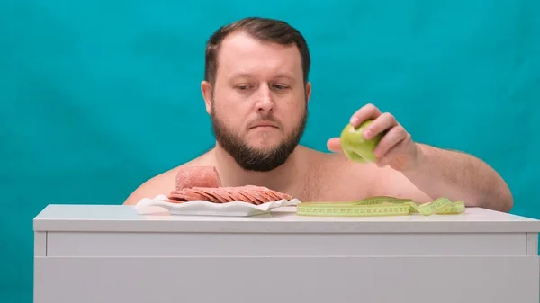 Bonito homem gordo faz uma escolha fácil entre uma maçã verde ou salsicha, escolhendo frutas orgânicas saudáveis em vez de fast food insalubre, fechando os olhos e sorrindo alegremente — Fotografia de Stock