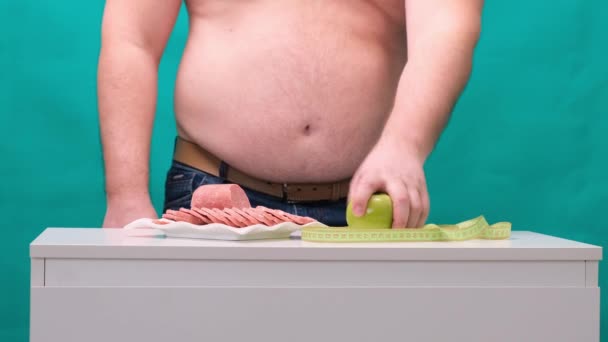 Человек с толстым животом выбирает между колбасой и зеленым яблоком. Концепция диеты и силы воли . — стоковое видео