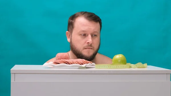 Homem gordo barbudo escolhe entre uma salsicha ou uma maçã verde em uma tela verde. Conceito Resistência à tentação, fast food, alimentação saudável, dieta, cuidados com o corpo . — Fotografia de Stock