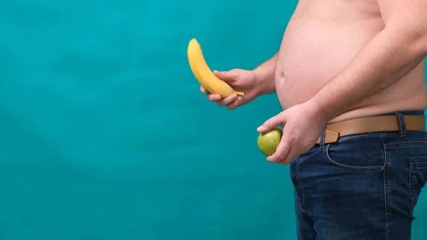 Hombre gordo con un vientre grande sostiene una manzana verde y un plátano en sus manos. El concepto de alimentación saludable y pérdida de peso, dieta . — Foto de Stock
