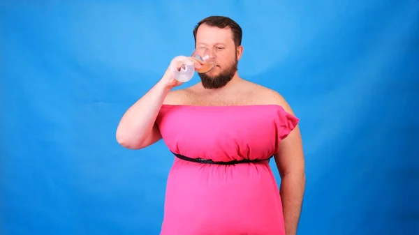 Забавный бородатый парень в розовом платье из подушек пьет вино из стакана на голубом фоне. Сумасшедший карантин. Мода 2020. Положи подушку. Вызов 2020 из-за изоляции дома — стоковое фото