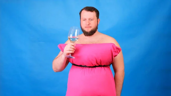 穿着睡椅做的粉红连衣裙，留着有趣胡子的家伙，喝着蓝色背景杯子里的酒。疯狂的检疫。时尚2020 。穿上枕头。2020年住房隔离带来的挑战 — 图库照片