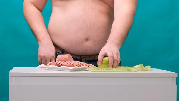 El hombre con el vientre gordo elige entre la salchicha y la manzana verde. El concepto de dieta y fuerza de voluntad . — Foto de Stock