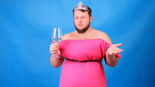 Divertente ragazzo barbuto in un abito rosa fatto di cuscini beve vino da un bicchiere su uno sfondo blu. Una quarantena pazzesca. Moda 2020. Mettiti un cuscino. Sfida 2020 a causa dell'isolamento della casa — Foto Stock