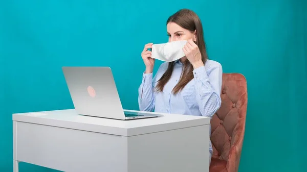 Kobieta zakłada maskę ochronną podczas pracy na laptopie w miejscu pracy lub w domu podczas pandemii. Koncepcja pracy podczas kwarantanny i samoizolacji. — Zdjęcie stockowe