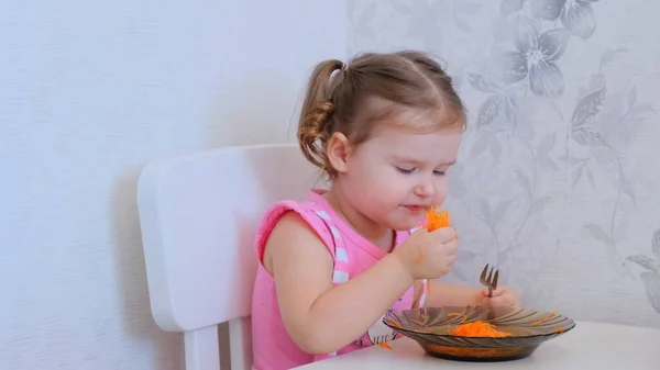 Bella ragazza per due anni mangiare carote grattugiate a un piccolo tavolo bianco e indulge. Concetto nutrizionale sano per i bambini — Foto Stock