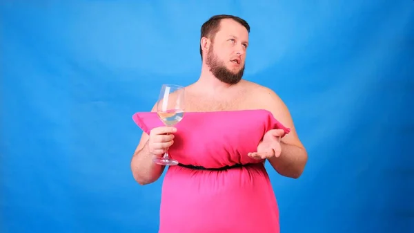 Rolig skäggig kille i rosa klänning gjord av kuddar dricker vin från ett glas på en blå bakgrund. Galen karantän. Mode 2020. Sätt på dig en kudde. Utmaning 2020 på grund av isolering — Stockfoto