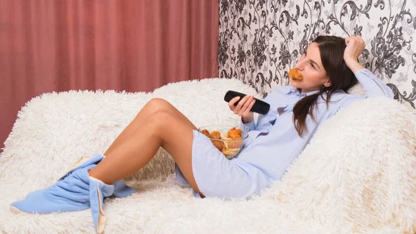 beautiful woman is bored in front of the TV, switches channels and eats junk food chicken wings. The concept of quarantine at home, idleness, unhealthy diet
