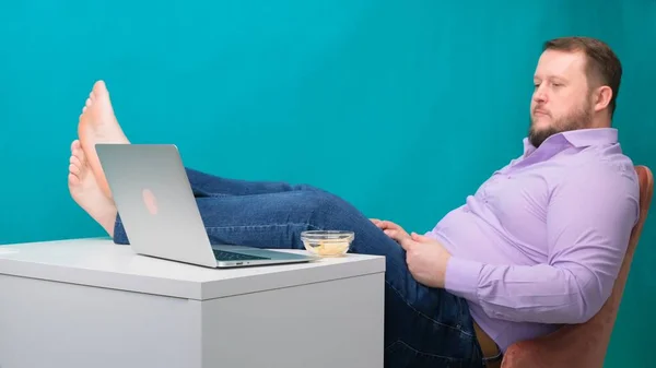 Потрібен час, щоб розслабитися. Розслаблений молодий чоловік тримає ноги на столі, працює за комп'ютером і їсть чіпси — стокове фото