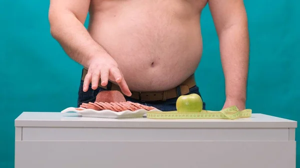 Mann mit dickem Bauch wählt zwischen Wurst und grünem Apfel. Das Konzept der Ernährung und Willenskraft. — Stockfoto