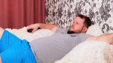 Sakallı yakışıklı şişman adam televizyon izliyor uzaktan kumandayı kullanarak kanal değiştiriyor. Erkek TV ekranında gördüklerinden bıkmış durumda. Evdeki oturma odasındaki kanepede uzanıp, fotokopi çekmek..