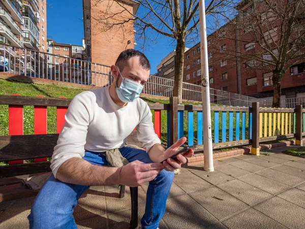 Parkta telefonuna bakan genç bir adam kendini Coperid-19 Coronavirus 'tan korumak için maske takıyor. Kavram salgını.