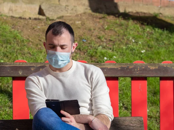 Parkta telefonuna bakan genç bir adam kendini Coperid-19 Coronavirus 'tan korumak için maske takıyor. Kavram salgını.