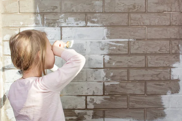 A little girl will paint a brick wall white. Wall painting, repairs in the house. The painter, the plasterer paints a wall. Finishing of facades with brick. Construction and repair work. Brickwork.