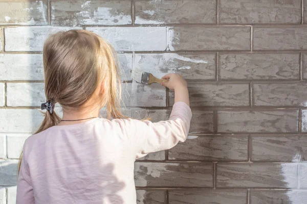 The painter, the plasterer paints a wall. Finishing of facades with brick. Construction and repair work. Brickwork. A little girl will paint a brick wall white. Wall painting, repairs in the house.