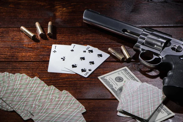 Gambling in poker or blackjack for money. On a wooden table are a pistol with cartridges, cash dollars, and a deck of playing cards with an ACE of spades and a six. Dangerous gambling debts and loans