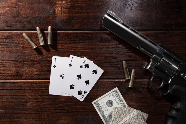 Gambling in poker or blackjack for money. On a wooden table are a pistol with cartridges, cash dollars, and a deck of playing cards with an ACE of spades and a six. Dangerous gambling debts and loans