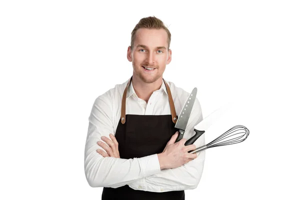 Knappe lachende chef-kok in wit en zwart, bedrijf 2 messen en een vliegenmepper kijken camera staan tegen een witte achtergrond. — Stockfoto