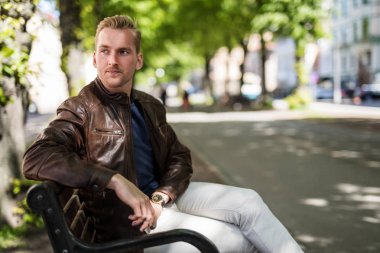 Rahat genç adam açık havada uzağa arıyorsunuz bir kahverengi deri ceket giyen bir bankta oturuyor. Güneşli yaz gün.