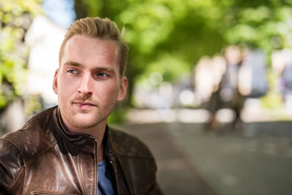 Relaxed young man sitting down on a bench outdoors wearing a brown leather jacket looking away. Sunny summer day.