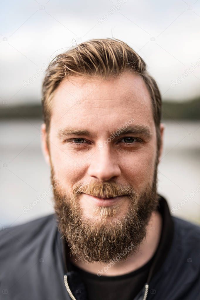 Laid back man with beard standing against a big lake looking at camera outdoors on a sunny day.