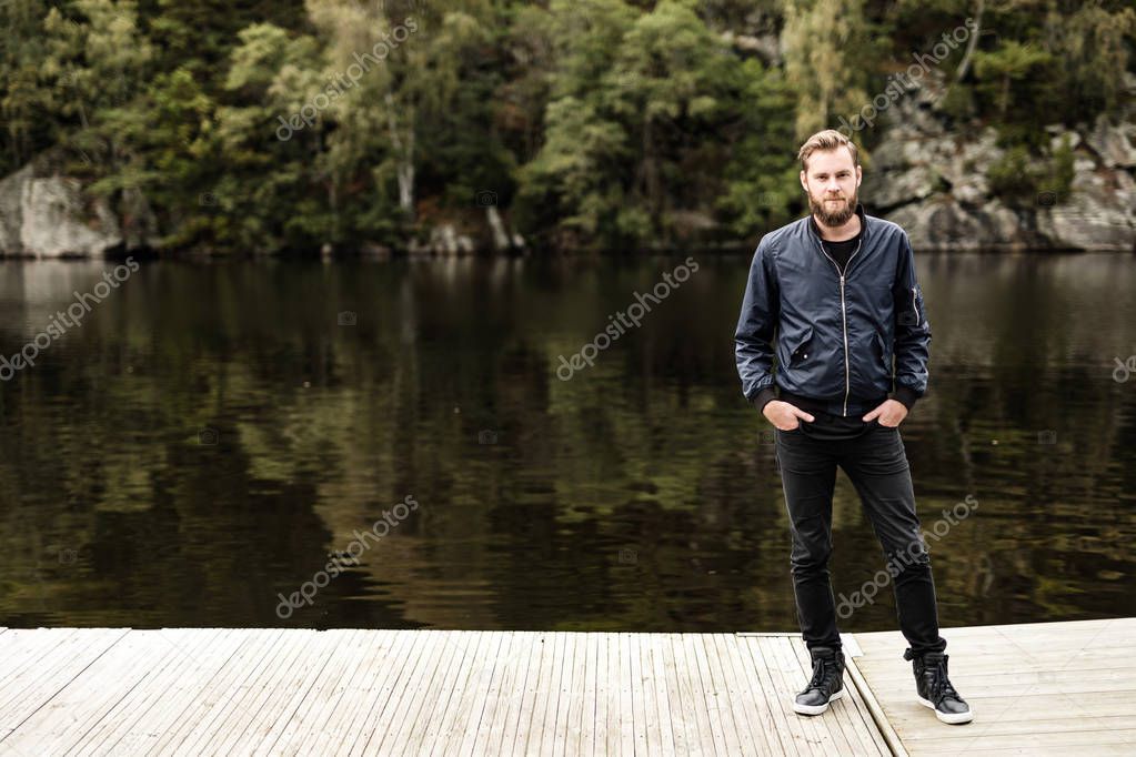 One bearded man wearing a blue jacket, standing in front of a lake and trees on a moody day.