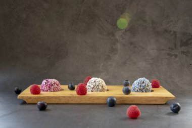 Üç farklı kalorili ev yapımı vegan enerji topları, ahşap tepside, koyu gri arka plan, ahududu ve yaban mersini.