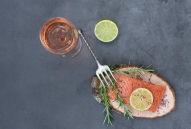 Bir dilim limonlu çiğ somon. Ağaç parçasında balık çatalı ve arka planda bir bardak gül şarabı var.