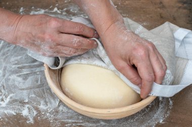 Mutfak havlusuyla kaplanmış iki el ekmek hamuru tahta masanın üzerindeki yalıtım sepetinde yükseliyor.