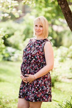 Çiçek desenli hamile kadın hamile elbisesi içinde parkta rüya gibi görünüyor. Doğal yaşam tarzı görüntüsü Mayıs 'ta çekildi. Son üç aylık dönemindeki bir kadın. Parkta hayat tarzı çekimi. Doğal makyaj yok bakışı. Sarışın, kısa saçlı. Bulanık arkaplan.