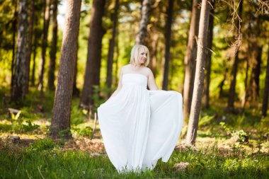 Güneşli bir yaz gününde, ormanın ortasında beyaz hamile bir kadının portresi rüya gibi görünüyor. Göbeğini tutan sarı saçlı çekici bir kadının portresi.. 