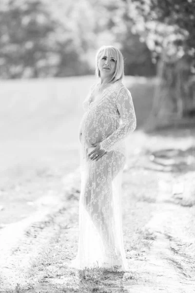 Portrait of a beautiful pregnant woman in sheer long white maternity dress looking dreamy in the middle of a forest on a sunny summer day. Portrait of an attractive lady with blond hair holding her belly.