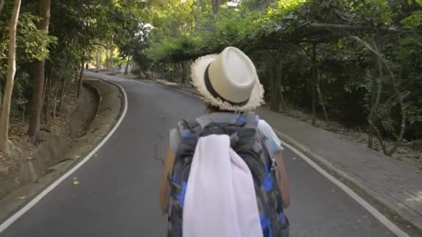 熱帯雨林をバックパックで歩くアジアの女性ハイカーの背面ビュー 藁帽子をかぶった女性が丘の上を目指して小さな道を歩いている — ストック動画