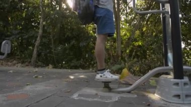 Sırt çantalı Asyalı kadın spor ekipmanı kullanıyor ve halka açık bir parkta güneş ışığı altında cep telefonu kullanıyor..