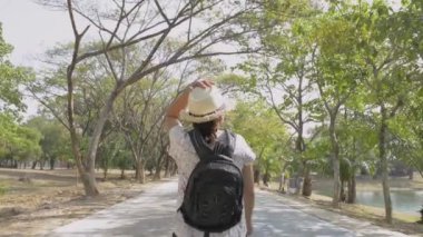 Sırt çantalı ve hasır şapkalı Asyalı kadının arkadan görünüşü yaz boyunca halka açık parkta yürüyor..