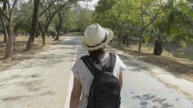 Sırt çantalı ve hasır şapkalı Asyalı kadının arkadan görünüşü yaz boyunca halka açık parkta yürüyor..
