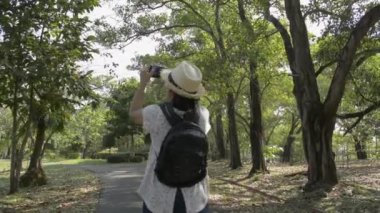 Sırt çantalı ve hasır şapkalı Asyalı kadının arkadan görünüşü yaz boyunca halka açık parkta cep telefonuyla yürüyüp fotoğraf çekmek..